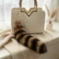 Brown-and-black fluffy animal-tail bag charm with a gold-tone clasp, shown on a cream handbag - Raccoon Tail / 30cm