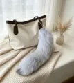 Gray fluffy tail bag charm with gold-tone hook and chain, displayed on a cream tote bag - Blue Fox Tail