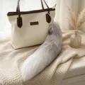 Gray-white gradient fox-tail plush bag charm with a gold clasp, hanging beside a cream handbag - Cross Fox Tail