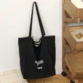 Bolso tote negro de lona con asas largas y letras blancas, colgado junto a una estantería de madera - 46*14*37 / Negro