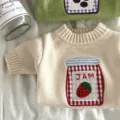 Detalle del tejido crema, cuello acanalado y parche de cuadros con un tarro de mermelada de fresa - XS / Beige