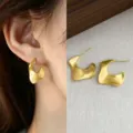 Close-up of an ear wearing a matte brushed gold curved earring beside a pair on textured fabric - Irregular Brushed Earrings (5304)