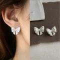 Close-up of a silver-tone pleated bow stud worn on the ear, with its pair displayed on linen. - Glazed Bowknot (6124) / Silver
