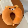 Close-up of a plush orange bear-shaped neck pillow with a white snap button on a wooden table - Bear / Orange