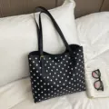 Bolso negro de lunares blancos, cierre dorado y asas largas, sobre una cama junto a un libro abierto - Negro