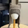 Chunky ribbed knit texture and braided earflap ties on a gray-brown hat shown on a white mannequin. - Adult 54-60 One Size / Coffee
