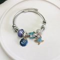 Pulsera plateada con colgantes de concha azul y estrella de mar en plato de cerámica blanca - Azul