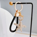 Close-up of golden starfish keychain with rhinestones and black braided leather strap - Starfish + Heart Strap / White