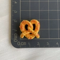 Dettaglio di un ciondolo a forma di pretzel in resina arancione di 2,5x2cm su scala graduata - Pane a Rotolo a Cuore