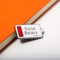 Pin esmaltado Social Battery en blanco y gris en el borde de un cuaderno naranja - Broche de Batería Blanco / X00426 - Actitud Social