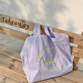 Detail of lavender canvas tote bag with yellow print resting on a wooden park bench - Purple