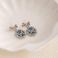 Detalle macro de pendientes de lazo plateados con patrón de mariposa sobre tela vaquera azul - Mariposa Ovalada