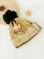 Metallic gold knit beanie with large black pompom on a cozy white plush background - One Size / Gold