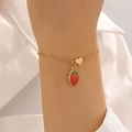 Dettaglio di un bracciale dorato con fragola smaltata rossa e ciondoli a cuore sul polso - Bracciale Stile Fragola (12382)