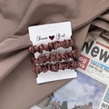 Three-pack of brown satin scrunchies displayed on silk fabric and newspaper backdrop - Rust red