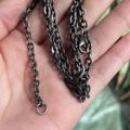 Close-up of black metal Figaro chain with lobster clasp held in hand against green background - Vintage Color Chain
