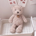 Brown curly plush teddy bear doll with a leather tag sitting in a cozy white storage basket - Light Brown Bear