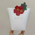Detail of white textured paper gift bag with red floral accent and gold merci print - Card + Two-legged Pin