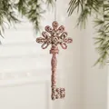 Pink gold glittery key-shaped Christmas ornament hanging on a festive green pine branch - Key Style