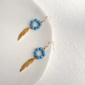 Dettaglio di orecchini boho con perline turchesi e pendenti a piuma dorata su piatto - 060 Piuma / Blu
