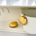 Dettaglio di orecchini a perno dorati lucidi esposti su una tazza bianca e una rivista - Grande Ovale Liscio / Oro