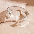 Detail of a shiny silver-tone fluid cuff bracelet resting on a textured stone surface - Silver Wave Texture Style / Silver