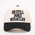Dettaglio del ricamo HE STILL DOES MIRACLES su un cappellino bicolore in cotone - Crema/Nero