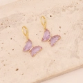 Detail view of gold butterfly earrings with purple and pink crystals on stone surface - Purple