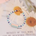 Detalle de pulsera de cuentas azules con colgante HAPPY sobre fondo de margaritas - Estilo HAPPY Rosa Verde