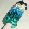 Stack of colorful silk satin scrunchies in blue, green, and neutral tones on a pedestal - Peacock Blue Gradient