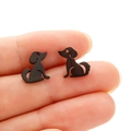 Detail view of small black stainless steel dachshund silhouette stud earrings on a finger - Sitting Dog (682) / Black