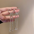 Detail view of rhinestone heart earrings with long tassels held against a beige background - Silver Needle Silver Heart Tassel Earring