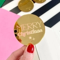 Golden round mirrored acrylic tag with Merry Christmas laser engraving held by hand - Gold Christmas