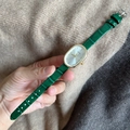 Detail of an oval gold watch with green leather strap held against a knit background - Green