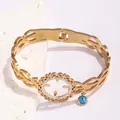 Gold hollow stainless steel bangle with diamond-set gear design and blue evil eye charm - Interwoven Hollow Style