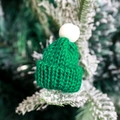 Mini green knit Christmas hat with white pompom on snow-covered pine branch - Green