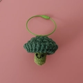 Handmade crochet broccoli pendant with smiling face and green wire loop on pink background - Broccoli