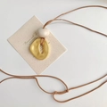 Lange braune Halskette mit unregelmäßigem Goldanhänger und weißer Perle auf beiger Karte - Braunes Seil Gold Unregelmäßiger Anhänger / Gold