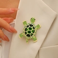 Elegante broche de tortuga dorada con gemas verdes en la solapa de una chaqueta crema - Verde