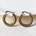 Détail de boucles d'oreilles créoles dorées avec pavé de pierres noires sur fond beige - Noir