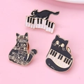 Tres broches de esmalte de gato y piano en negro, blanco y dorado sobre un fondo rosa suave - Juego de Broches Gato Piano Nota Musical