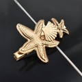 Detailed view of gold starfish and seashell hair clips with metallic texture on black background - Double Starfish Single Shell Duckbill Clip / Gold