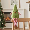 Niedliche Plüsch-Weihnachtsbaum-Puppe auf Holzstuhl vor einem warmen Kaminhintergrund - Grün