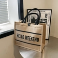 Beige jute tote bag with black handles and Hello Weekend text on a metal table - HELLO WEEKEND