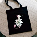 Black canvas tote bag featuring a pink letter J and elegant white lily print - Letter J / Black