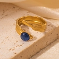 Detailansicht eines goldenen Federrings mit blauem Naturstein auf beigem Steinhintergrund - Blauer Stein Feder Stil (TYR0769)