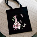 Black canvas tote bag featuring a pink letter E and watercolor lily flower print - Letter E / Black