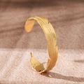 Detailed view of 18K gold plated stainless steel feather cuff bracelet on sandy texture - Feather
