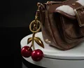 Close-up of cherry charm with red glitter on a brown monogram bag with white fleece trim - Red Cherry with Large Double Leaves (Bronze Gold)