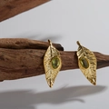Detailed close-up of leaf-shaped gold earrings with oval green peridot gemstones on wood - Earrings / Gold / Peridot